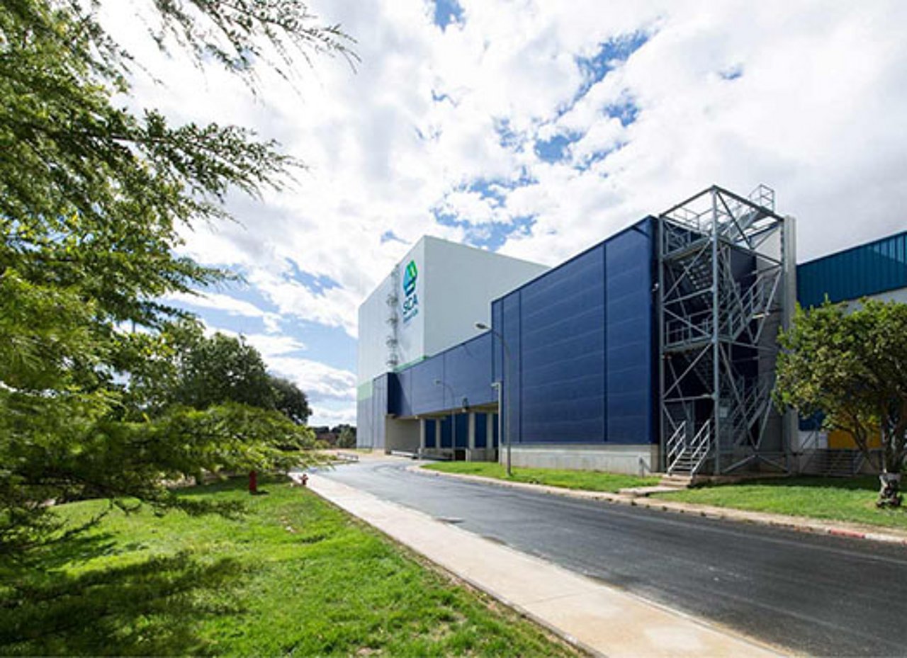 Man sieht ein Betriebshalle, davor eine Strasse und eine Wiese mit Bäumen.